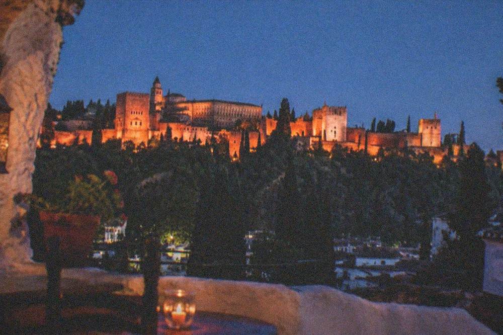 The view from a Sacromonte cave-house terrace at night, the floodlit Alhambra glowing red across the valley under a dark sky