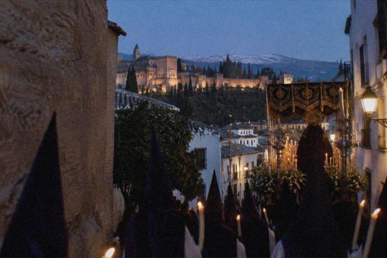 A candlelit religious procession moving through a narrow Granada street at night, with hooded penitents and a decorated float