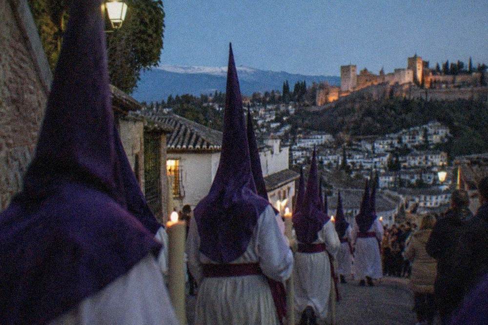 Hooded penitents in white and purple robes walking in formation at night, carrying long candles