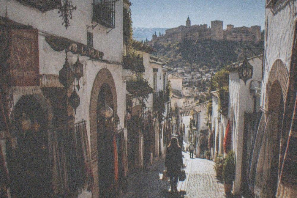A shopper carrying bags walking a cobbled street with the Alhambra visible on the hillside above