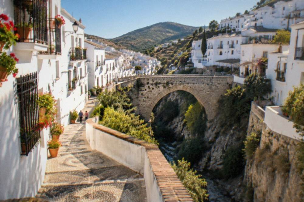 A stone bridge over a mountain gorge with a white village perched above it