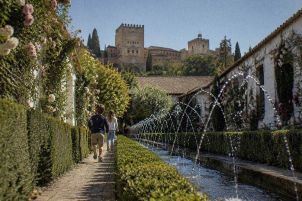 Children running along a path lined with hedges and fountains in the Generalife gardens