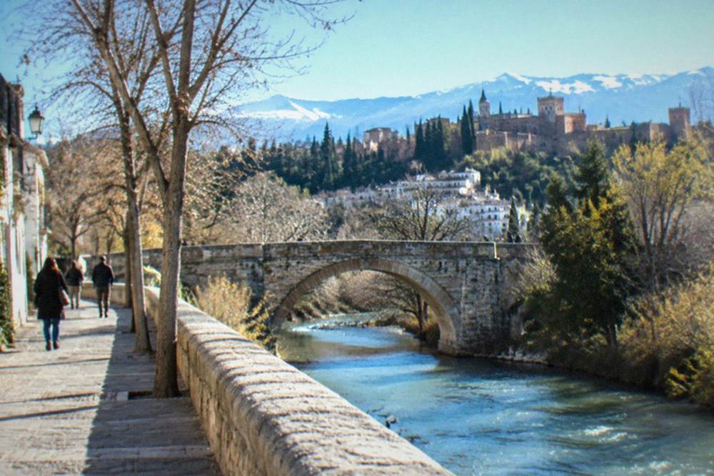 A flat riverside walk in Granada with an old stone bridge and trees along the water